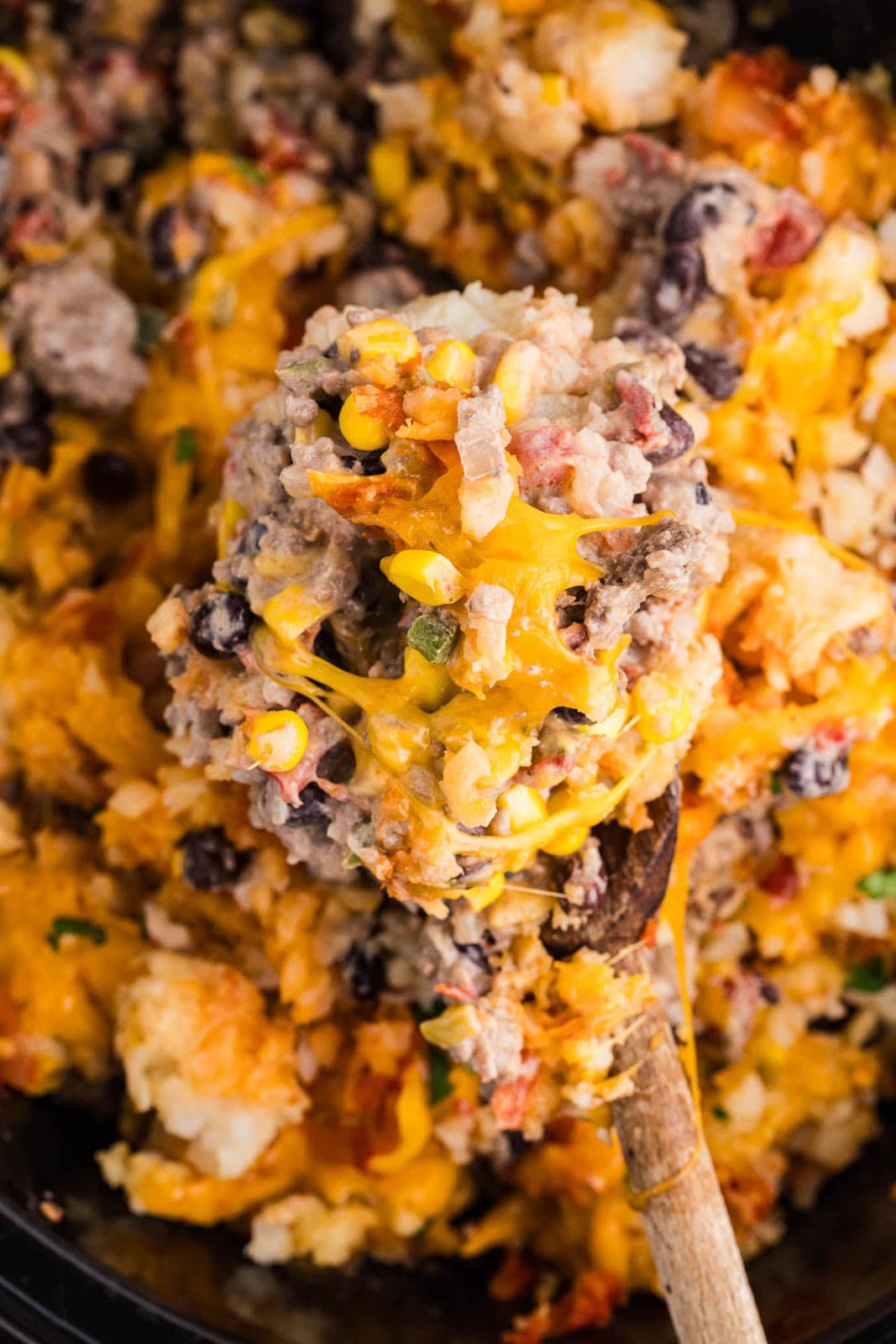 closeup of wooden spoon holding tater tots casserole with black slow cooker of casserole in background