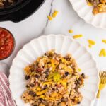 two white dinner plates of tater tot casserole, forks, salsa and black slow cooker in background