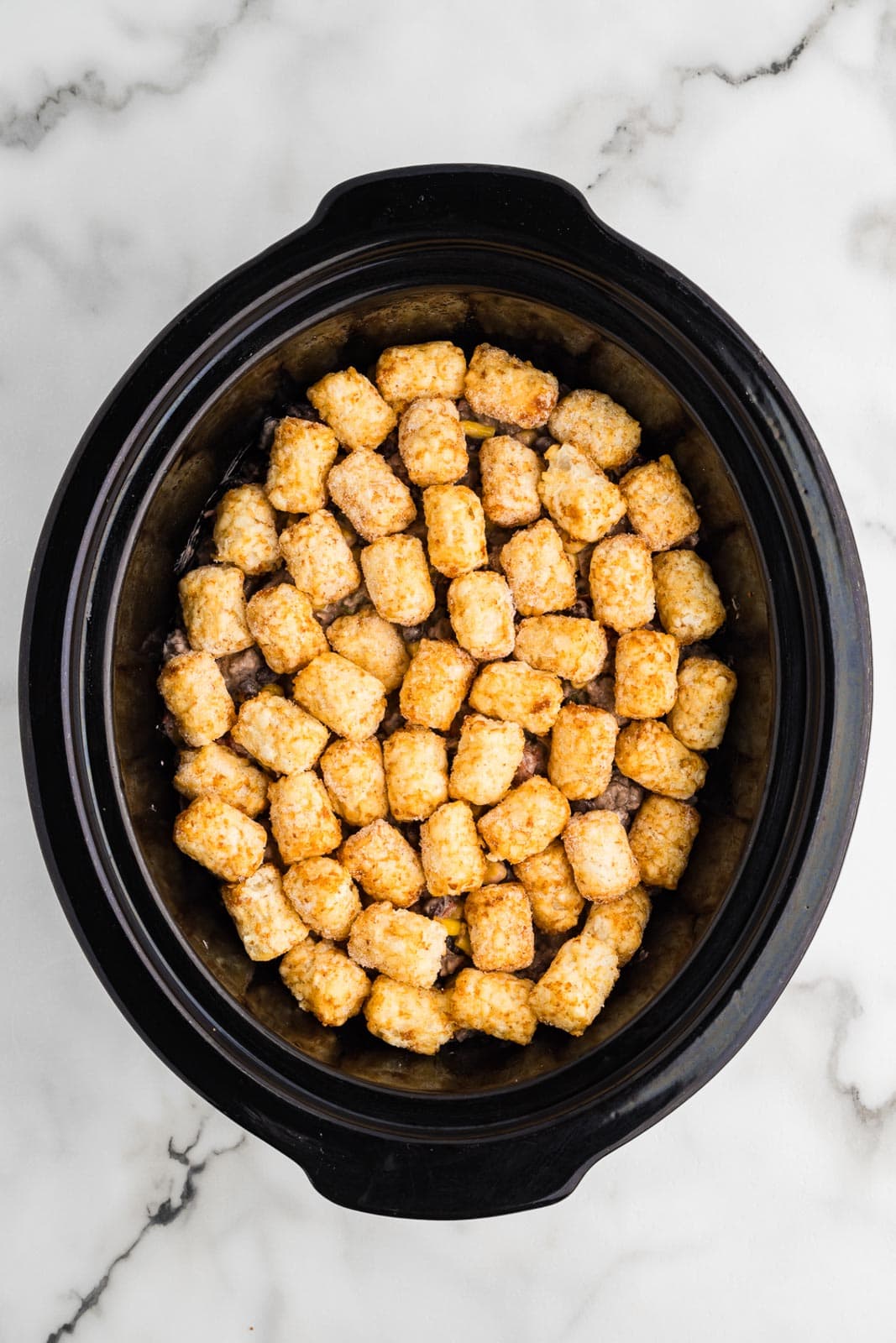 black slow cooker with tater tots, ground beef mixture and then topped with tater tots