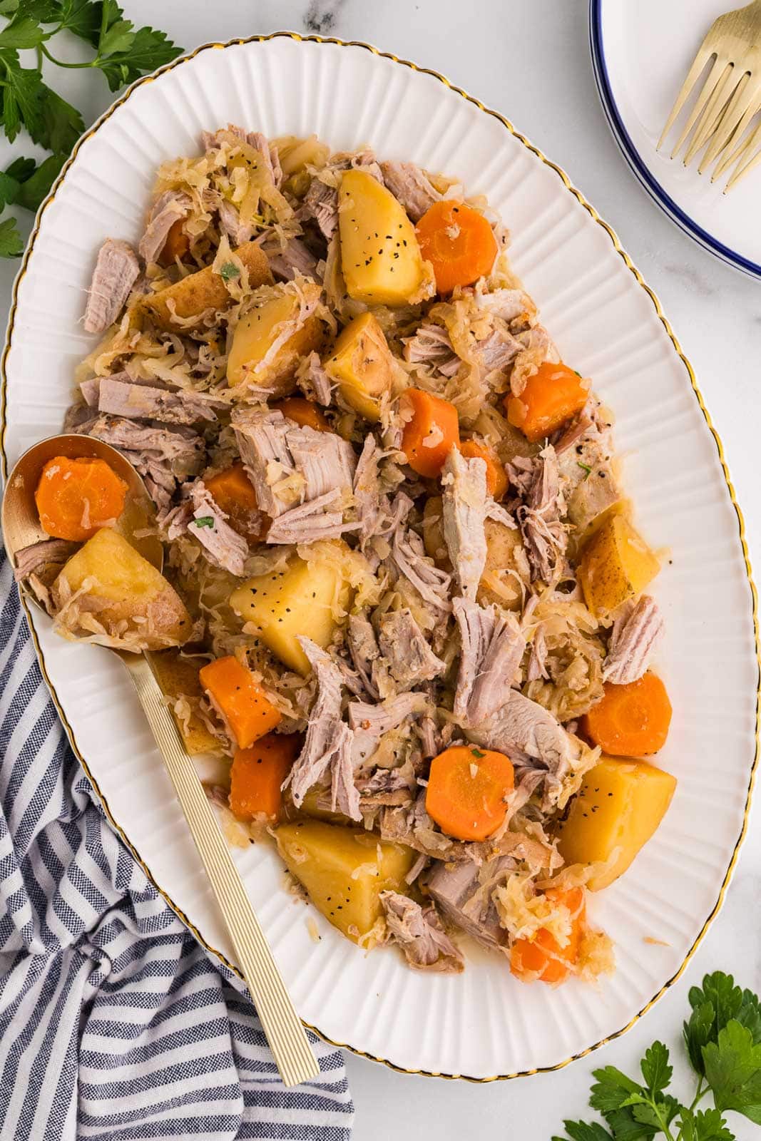 large white platter containing pork and sauerkraut with gold spoons, with another small plate and gold fork on side