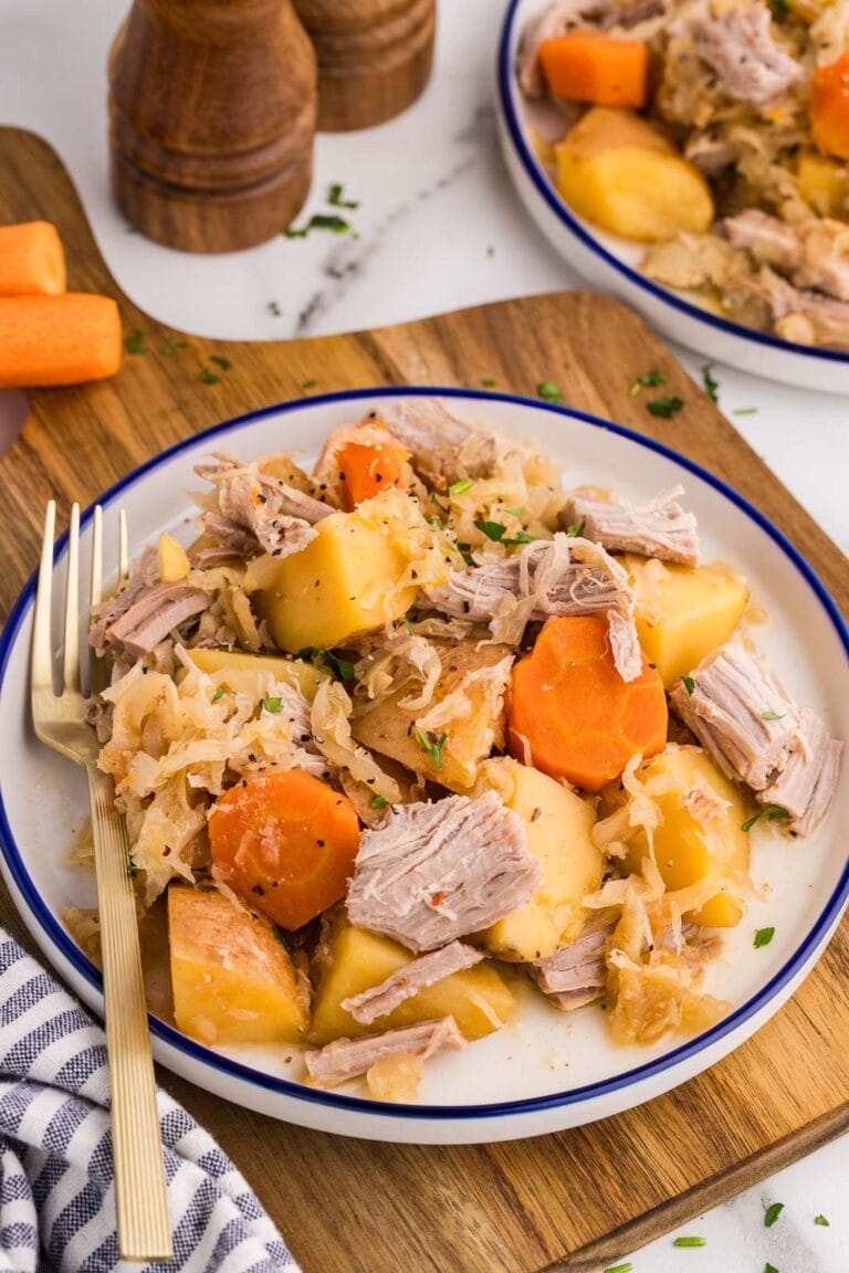 white dinner plate on wooden cutting board of pork and sauerkraut with gold fork, with another plate and salt and pepper shakers in background