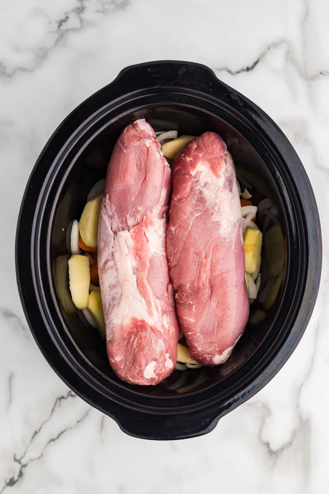 overhead view of black slow cooker with 2 pieces of pork loin atop vegetables