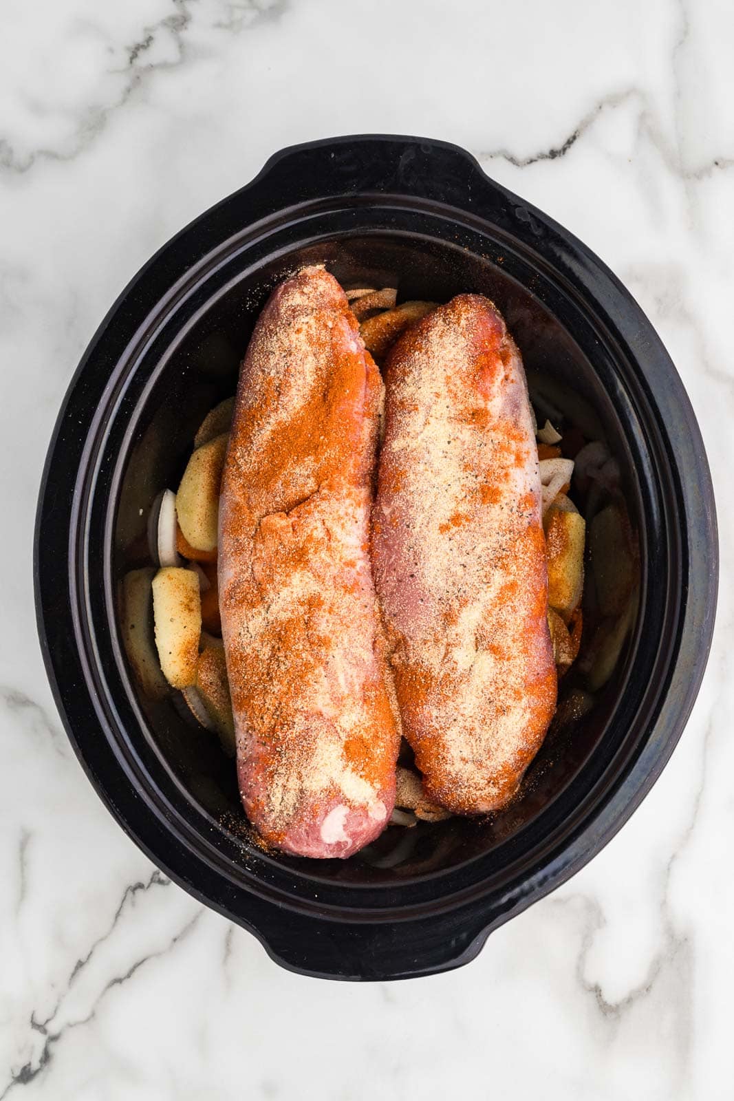 overhead view of black slow cooker with 2 pieces of seasoned pork loin atop vegetables