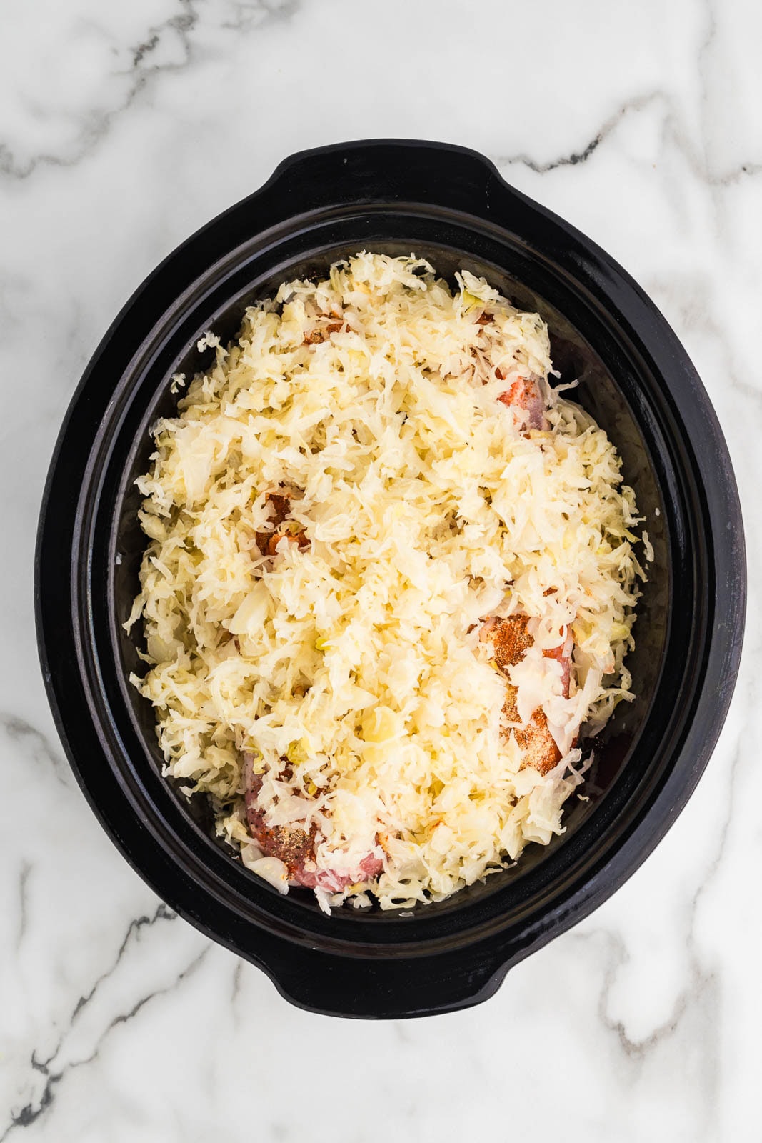 overhead view of black slow cooker with 2 pieces of seasoned pork loin atop vegetables with sauerkraut