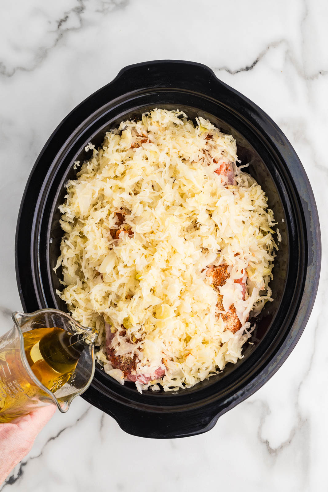 overhead view of black slow cooker with 2 pieces of seasoned pork loin atop vegetables with sauerkraut and hand pouring apple juice into slow cooker