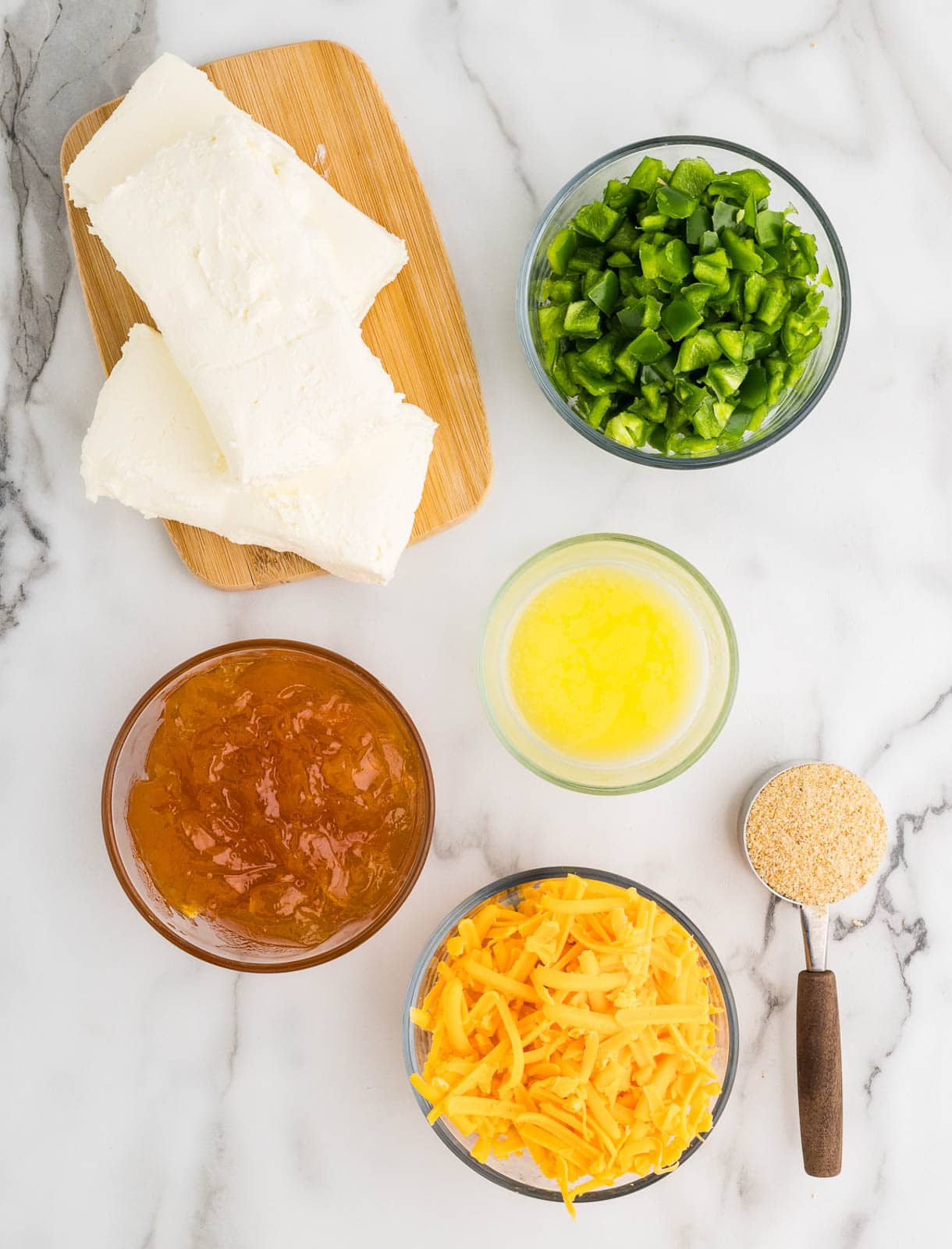 ingredients to include wooden cutting board with 3 blocks of cream cheese, surrounded by containers of diced jalapeno peppers, melted butter, shredded cheddar cheese, bread crumbs and apricot preserves