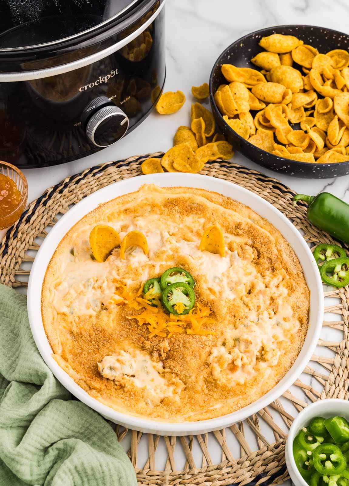 white serving bowl of jalapeno dip on trivet, surrounded by bowls or corn chips, preserves and jalapeno slices, alongside black slow cooker