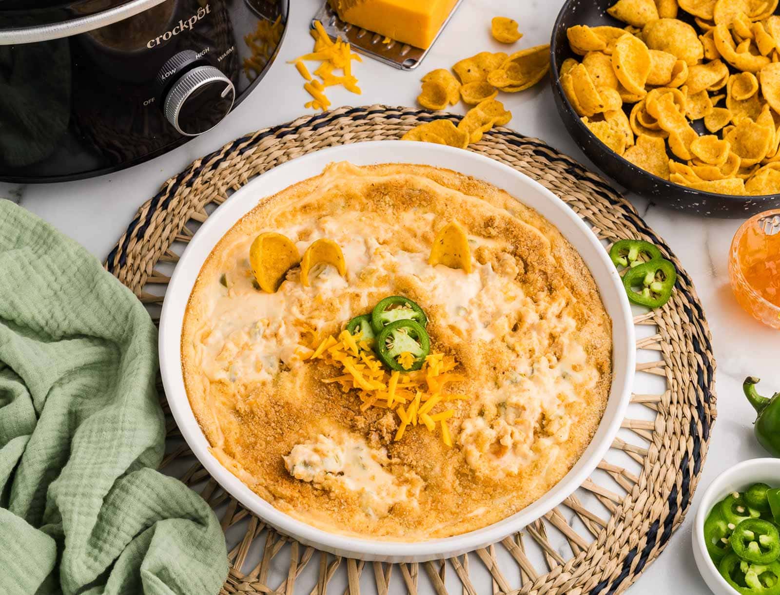 closer view of white serving bowl on trivet of dip, surrounded by bowls of corn chips, jalapeno slices and preserves, cheddar cheese with grater and black slow cooker