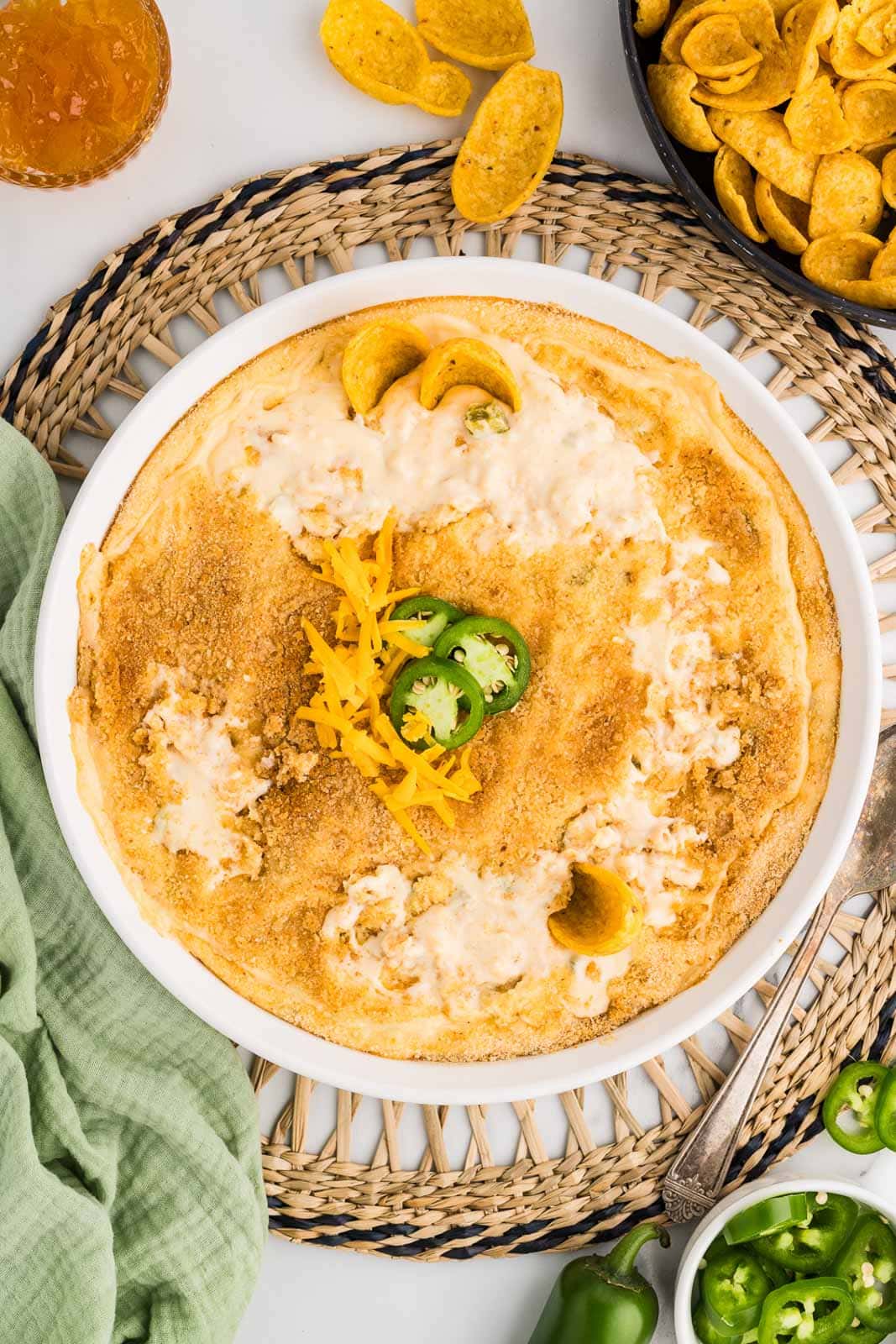 white serving bowl of jalapeno dip on a trivet, covered with bread crumbs, cheese, jalapeno pepper slices surrounded by bowl of corn chips, preserves, and jalapeno pepper slices