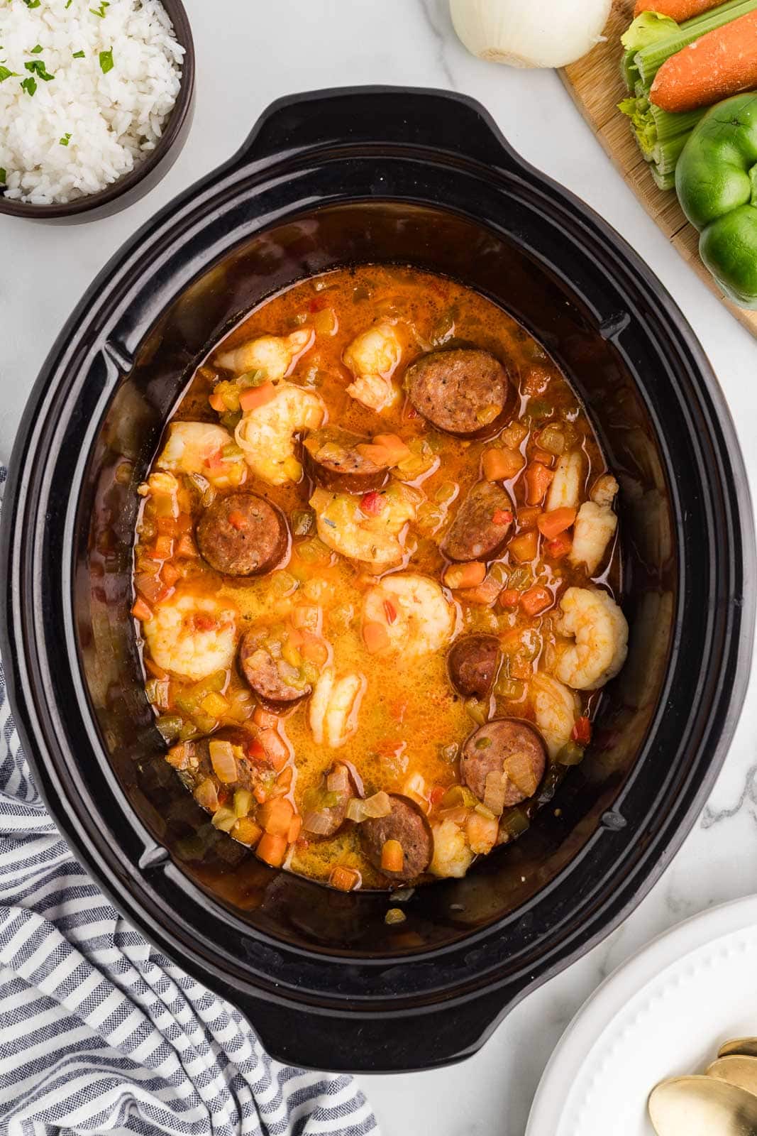 black slow cooker of prepare gumbo mixture surrounded by bowls of white rice and wooden cutting board with assorted whole vegetables