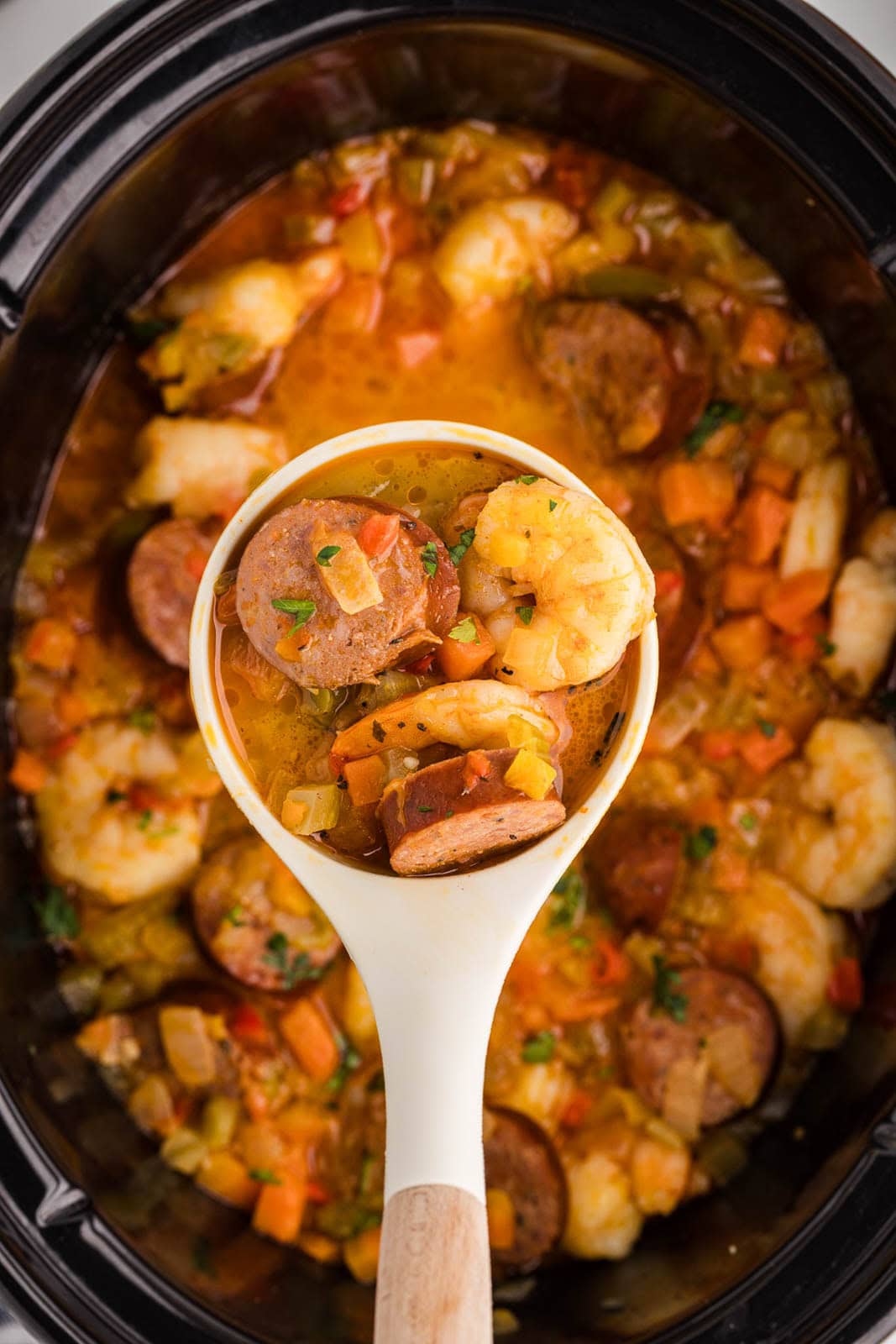 closeup of white ladle containing gumbo with black slow cooker of gumbo in background