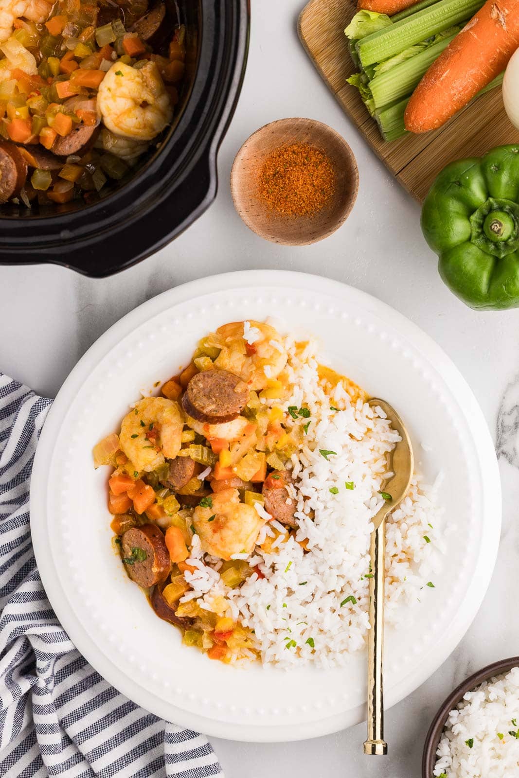 white bowl of gumbo and white rice with gold spoons, surrounded by black slow cooker of gumbo, bowls of seasoning and white rice, wooden cutting board of assorted raw vegetables