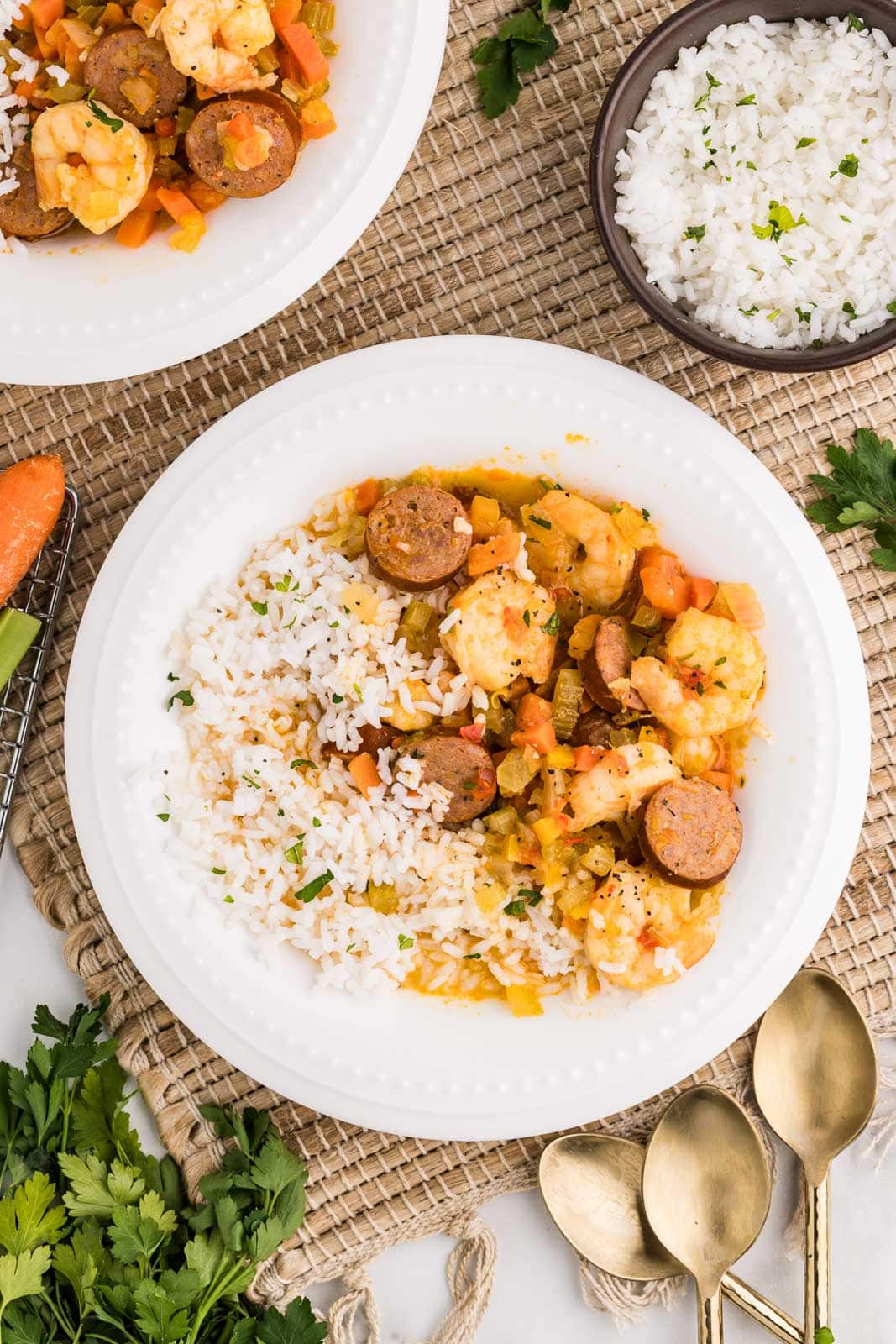 2 white bowls of gumbo and white rice with bowl of rice, 3 gold spoons and sprigs of parsley