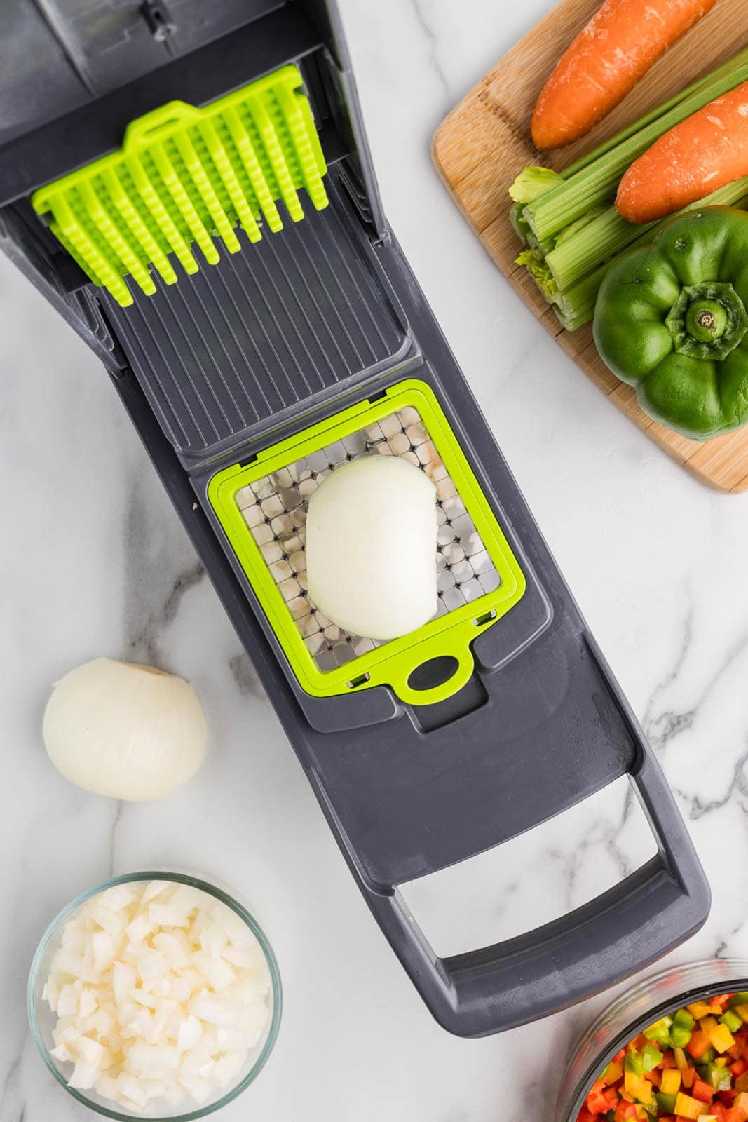 chopper with white onion on it surrounded by a wooden cutting board with green pepper, celery stalks and carrots, as well as bowls of diced peppers and white onion