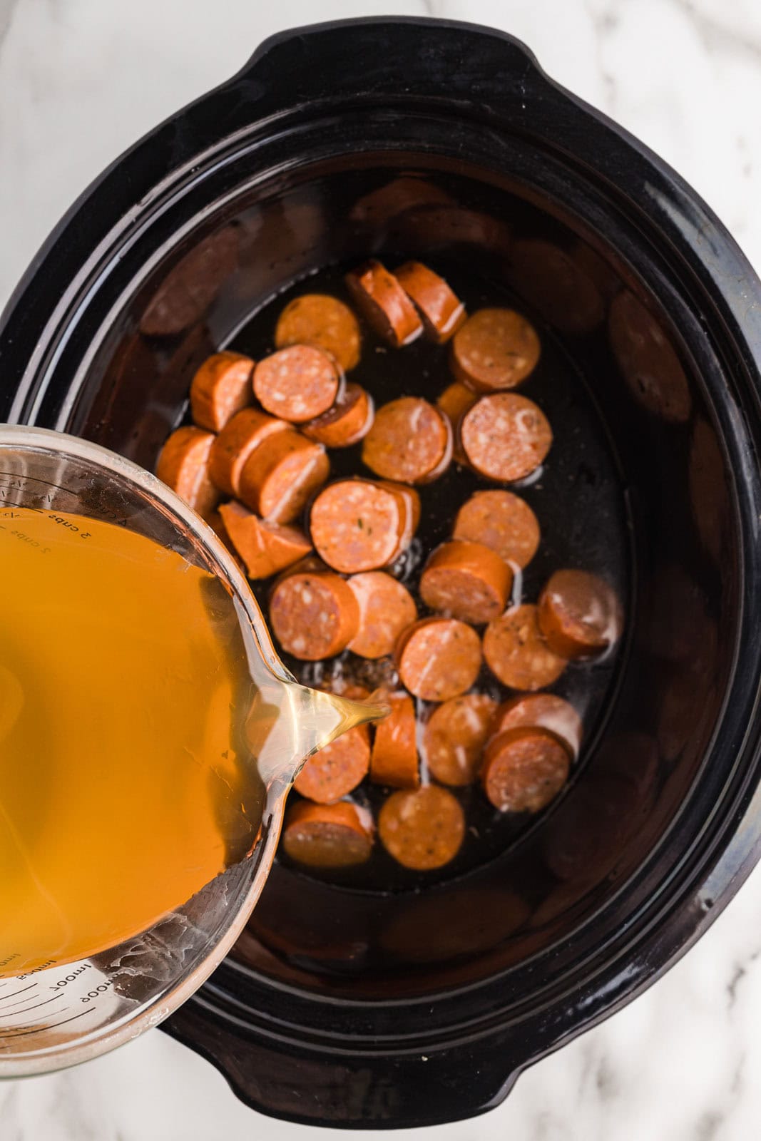 black slow cooker with sliced sausage with glass measuring cup of vegetable broth being poured into slow cooker