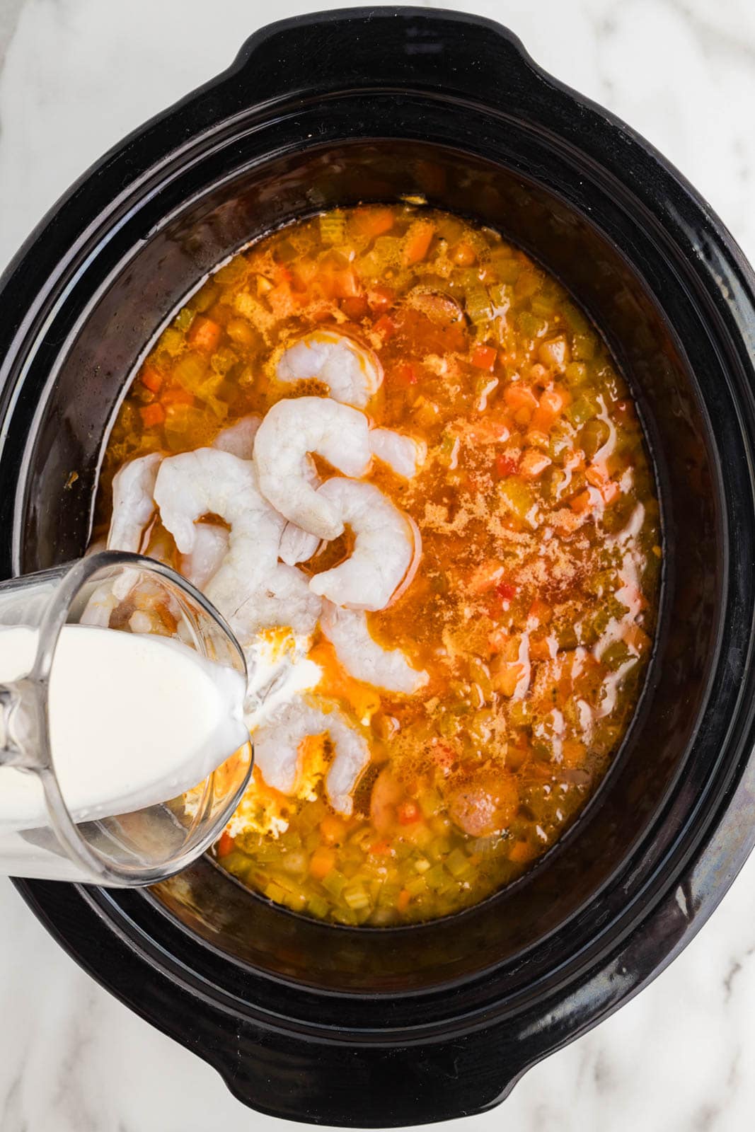 black slow cooker of vegetable mixture with shrimp added and cream being poured into slow cooker