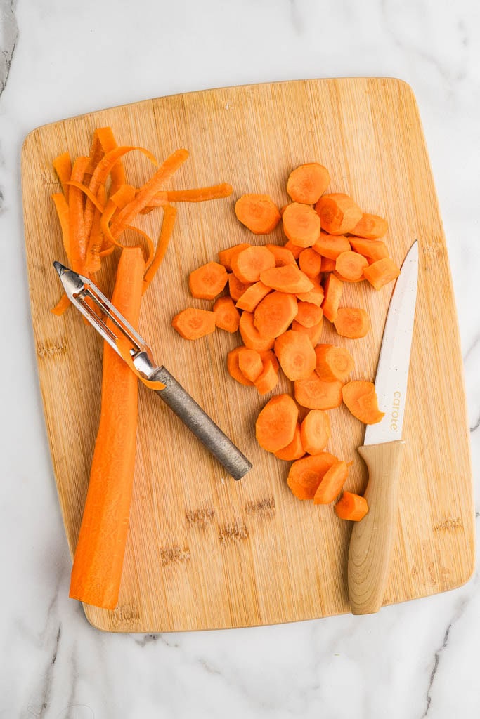 wooden cutting board with one whole peeled carrot, paring knife and peeler, cut up carrot and shaved pieces