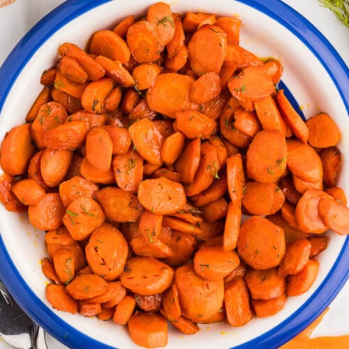 overhead view of sauteed carrots in white bowl with blue rim, with forks, salt, dill and carrots surrounding it