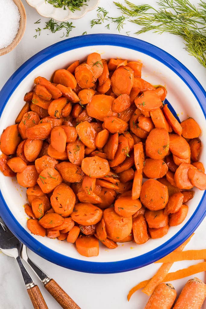 overhead view of sauteed carrots in white bowl with blue rim, with forks, salt, dill and carrots surrounding it