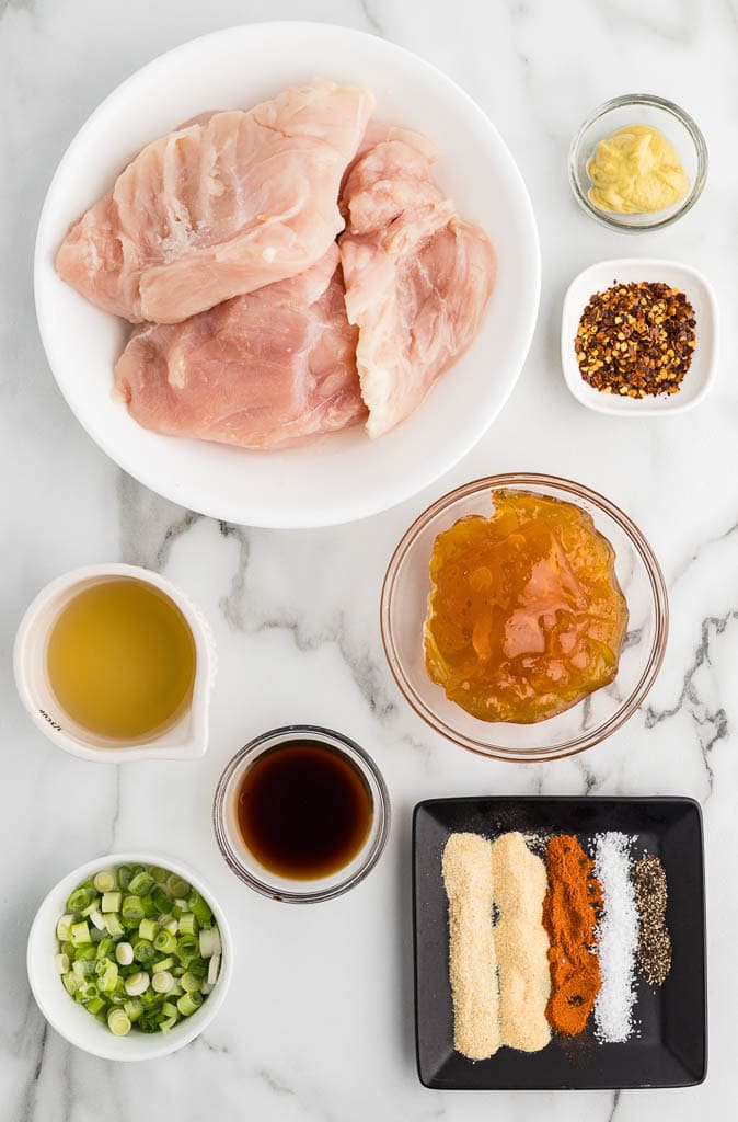 ingredients to include container of chicken breasts surrounded by containers of Dijon mustard, red pepper flakes, apricot preserves, chicken broth, soy sauce, sliced green onions and plate of garlic powder, onion powder, smoked paprika, salt and black pepper