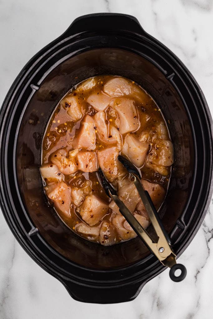overhead view of black slow cooker containing cubed chicken and all seasonings with tongs