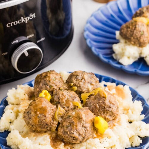 two blue dinner plate of mashed potatoes and meatballs with black slow cooker and salt and pepper mills in background