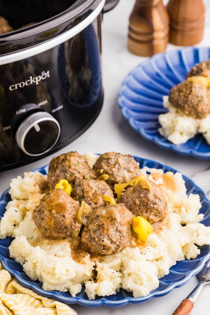 two blue dinner plate of mashed potatoes and meatballs with black slow cooker and salt and pepper mills in background