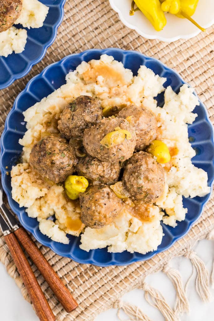 overhead view of blue dinner plate of mashed potatoes and meatballs on placemat with bowl of peppers, forks, and another dinner plate on side