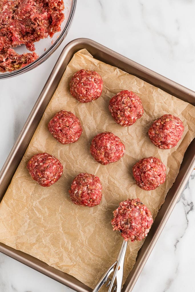 parchment paper lined cookie sheet with six meatballs with scoop, with glass bowl of meatball mixture in background