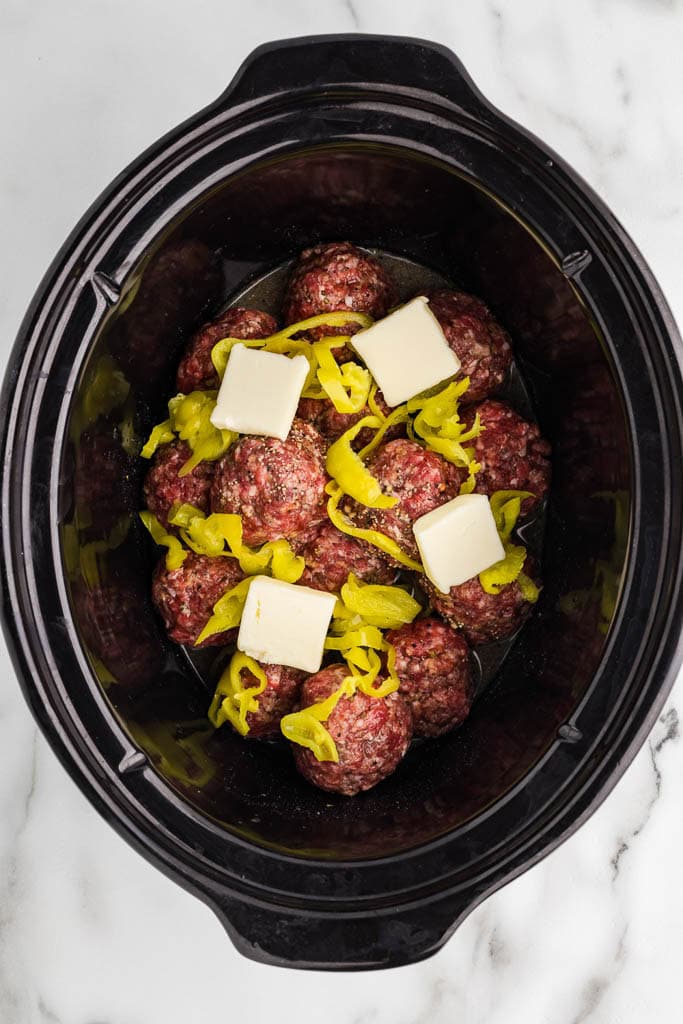 overhead view of black slow cooker containing meatballs with sliced peppers and pats of butter on top