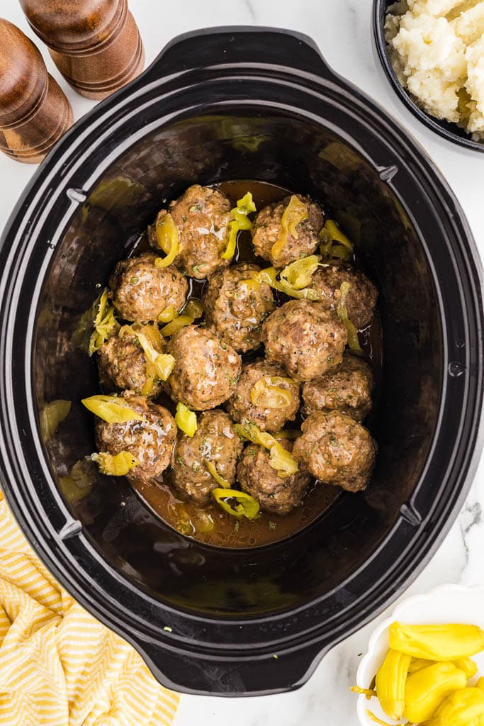 overhead view of black slow cooker with slow cooked meatballs surrounded by wooden salt and pepper mills, bowl of mashed potatoes, bowl of peppers and yellow dishtowel
