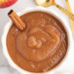 overhead view of white serving bowl with applesauce and a cinnamon stick inserted, surrounded by 2 gold spoons, an apple, another bowl of applesauce, and some cinnamon sticks
