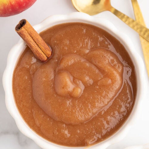 overhead view of white serving bowl with applesauce and a cinnamon stick inserted, surrounded by 2 gold spoons, an apple, another bowl of applesauce, and some cinnamon sticks