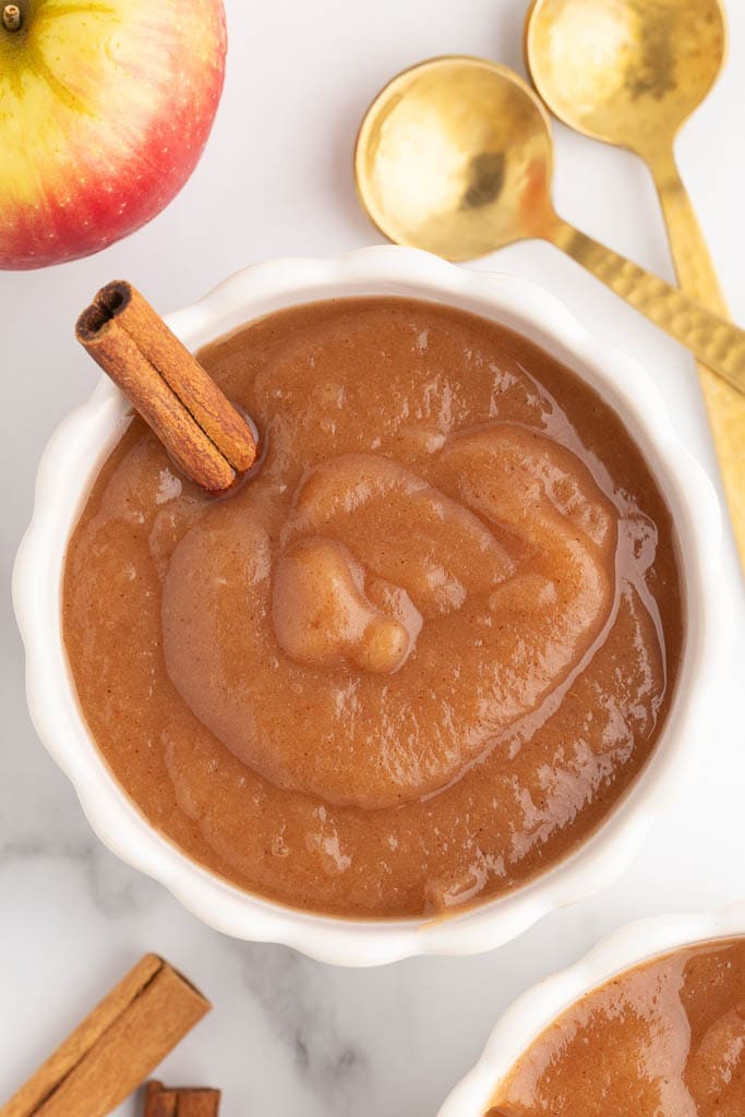 overhead view of white serving bowl with applesauce and a cinnamon stick inserted, surrounded by 2 gold spoons, an apple, another bowl of applesauce, and some cinnamon sticks