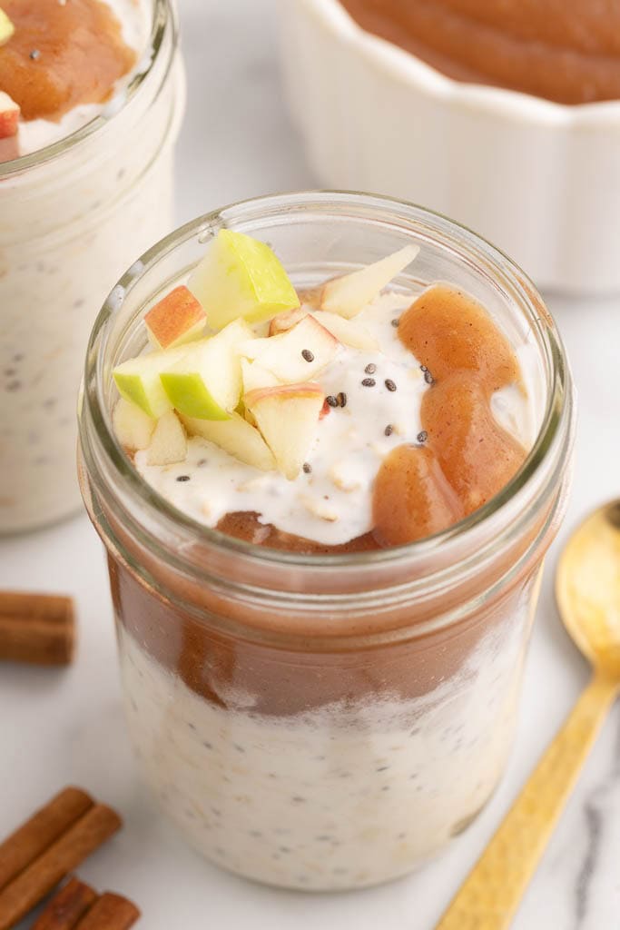 mason jars with yogurt topped with applesauce and raw apples, surrounded by another mason jar, gold spoons, cinnamon sticks and bowl of applesauce