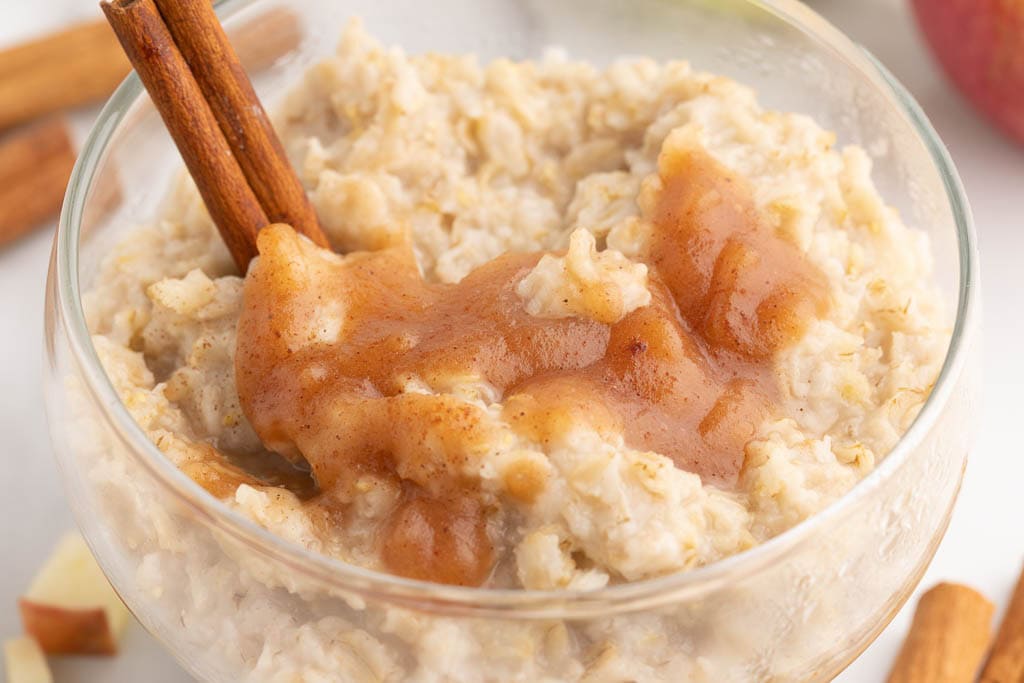 closeup view of glass bowl of oatmeal topped with applesauce and cinnamon sticks, surrounded by apples and cinnamon sticks in the background
