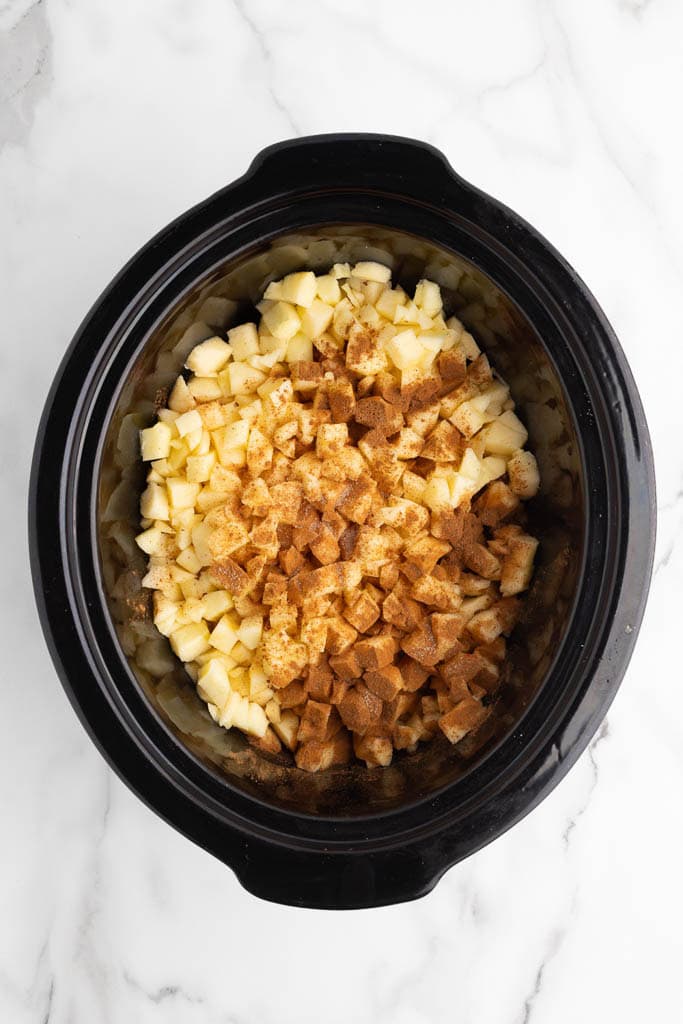 black slow cooker with peeled, cored and chopped apples sprinkled with cinnamon and all other ingredients