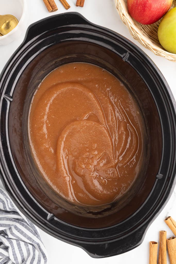 black slow cooker with cooked applesauce with basket of apples, and cinnamon sticks on side