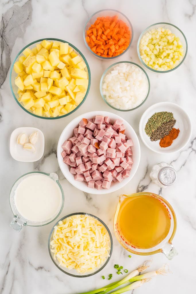 ingredients to include bowls of diced ham steak, diced potatoes, diced carrots, diced celery, diced onion, garlic cloves, chicken broth, seasonings, half and half, green onions, white cheddar cheese, and salt shaker