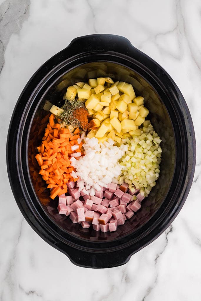 black slow cooker with ham, potatoes, carrots, celery, onion, garlic, broth, thyme, pepper, and smoked paprika