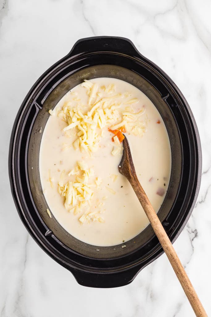 black slow cooker with soup ingredients with half and half and white cheddar cheese with wooden spoon