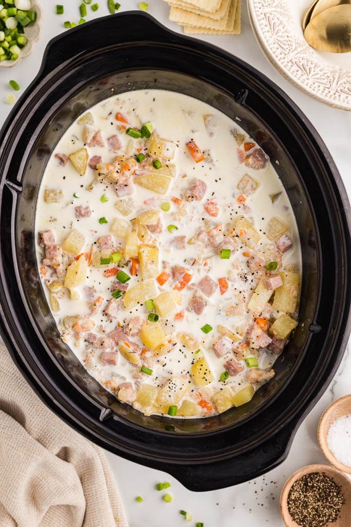 overhead view of black slow cooker with slow cooked soup surrounded by green onions, crackers, bowls with spoons, salt and pepper and tea towel