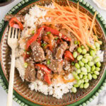 overhead view of dinner plate of teriyaki beef on white rice with edamame and carrot sticks and gold fork, surrounded by containers of cucumber, salt and pepper, sesame seeds, green onion, and carrots sticks