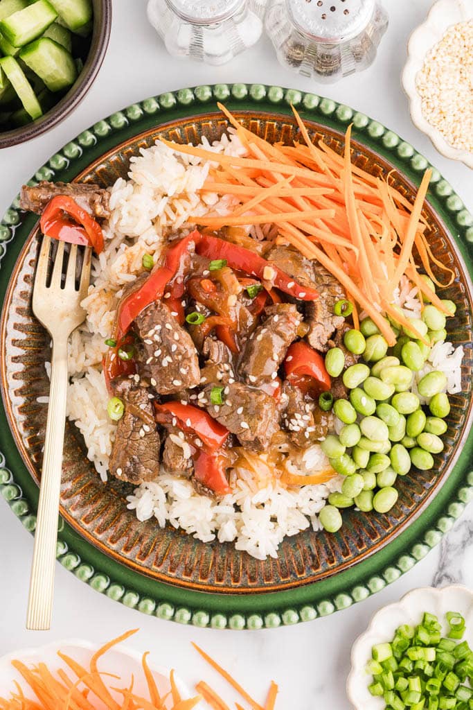 overhead view of dinner plate of teriyaki beef on white rice with edamame and carrot sticks and gold fork, surrounded by containers of cucumber, salt and pepper, sesame seeds, green onion, and carrots sticks