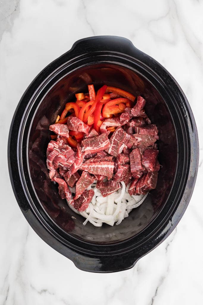 overhead view of black slow cooker onions, red peppers and sliced beef