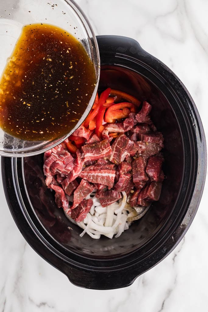 overhead view of black slow cooker onions, red pepper and beef slices with glass bowl of sauce being poured over