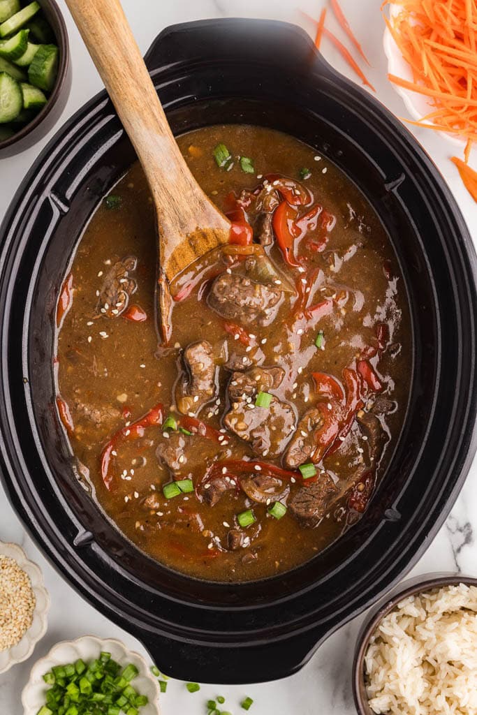 overhead view of black slow cooker containing slow cooked beef teriyaki with wooden spoon inserted, surrounded by bowls of cucumber, carrots, rice, green onions and sesame seeds